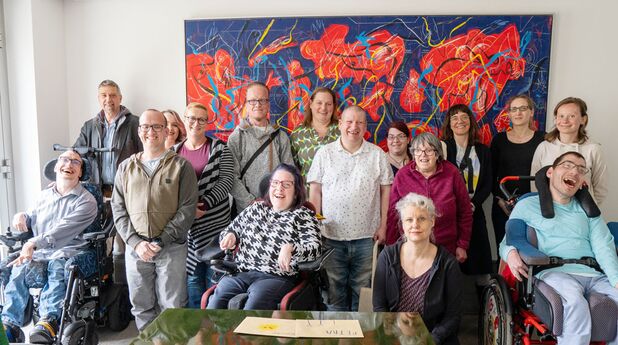 Ein Foto einer großen Personengruppe vor einem bunten Gemälde. Alle lache uns an. Einige der Personen sitzen in Rollstühlen.