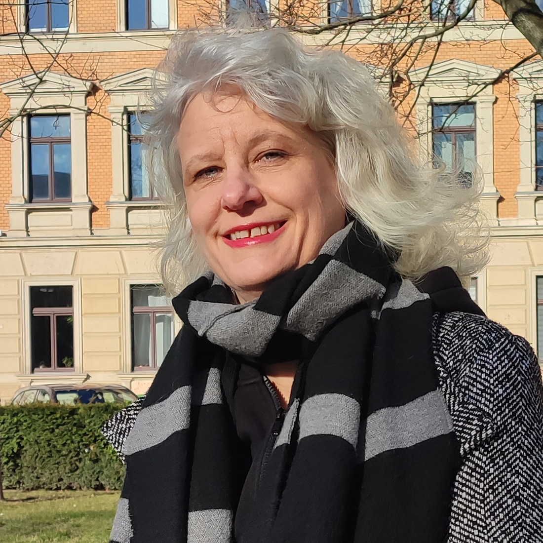 Eine Frau mittleren Alters lächelt mit offenem Mund in die Kamera. Sie hat graue, lockige, schulterlange Haare. Ihr Mund ist rot geschminkt. Sie trägt einen grau-schwarzen Schal und Mantel.